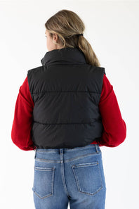 A woman viewed from behind wearing a black puffer vest over a red long-sleeve top and blue jeans, against a plain white studio background.