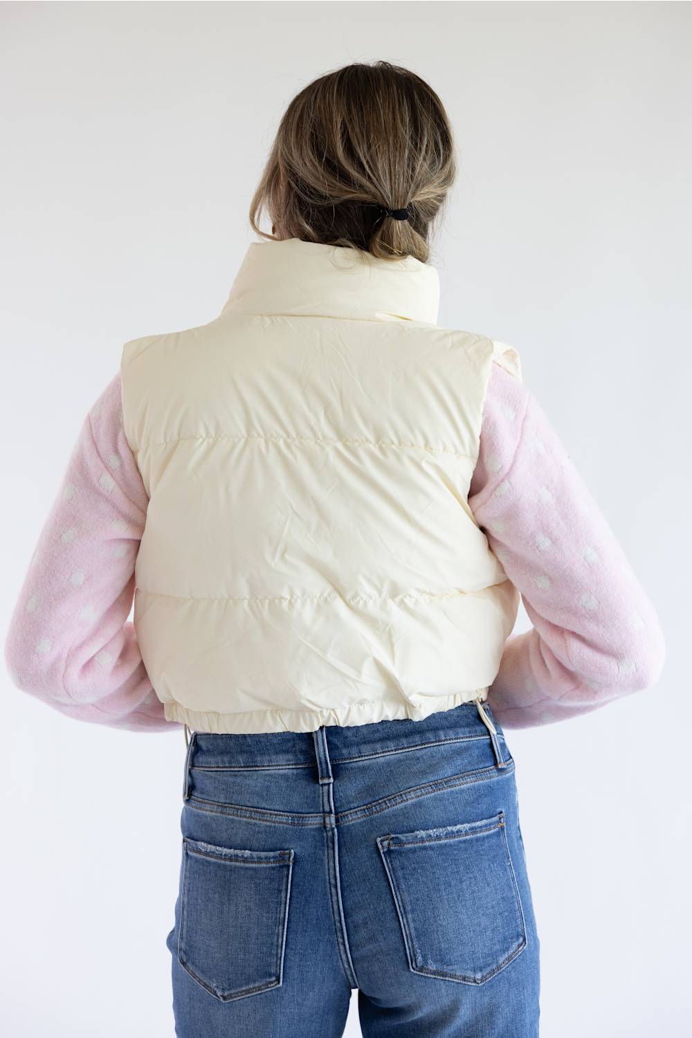A person shown from the back wearing a cream puffer vest over a light pink long-sleeve top and blue jeans, against a plain white background.