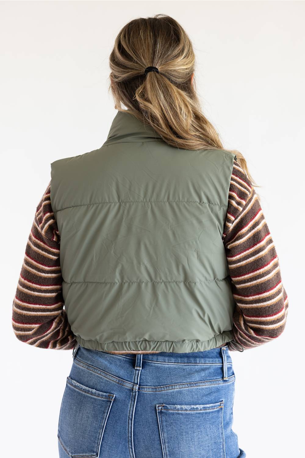 A woman shown from behind wearing an olive green puffer vest over a striped long-sleeve shirt and blue jeans, against a plain white background.