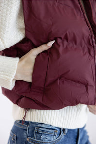 A close-up of a person wearing a quilted puffer vest in deep burgundy, styled over a cream knit sweater and blue jeans.