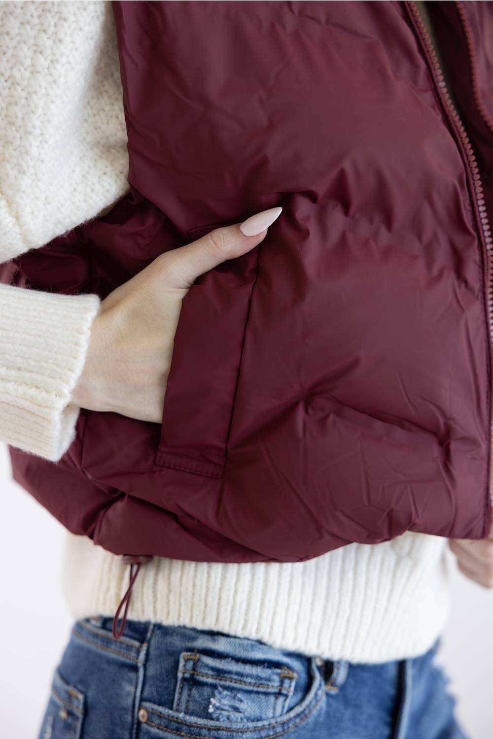 A close-up of a person wearing a quilted puffer vest in deep burgundy, styled over a cream knit sweater and blue jeans.