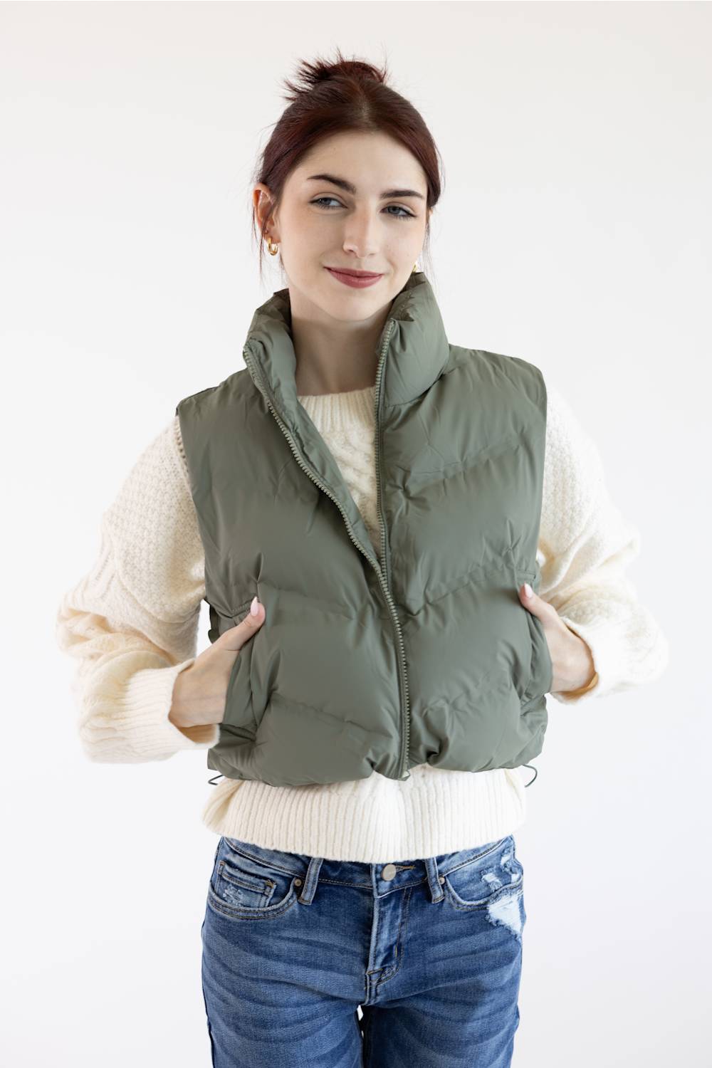 A woman wearing an olive green puffer vest over a cream knit sweater and blue jeans, standing against a plain white studio background.