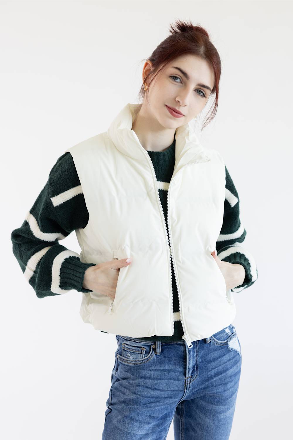 A woman wearing a cream-colored sleeveless puffer vest over a black and white striped knit sweater, paired with blue jeans, against a plain white background.