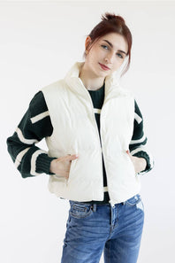 A woman wearing a cream-colored sleeveless puffer vest over a black and white striped knit sweater, paired with blue jeans, against a plain white background.