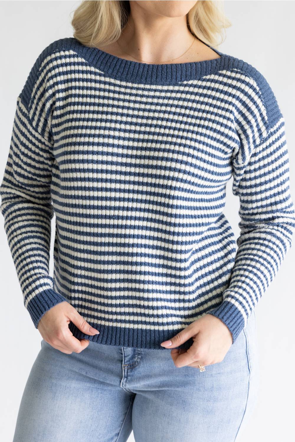 A woman wearing a blue and white horizontally striped knit sweater with a wide boat neckline, paired with light blue jeans, shown from the shoulders to the hips against a plain light gray background.