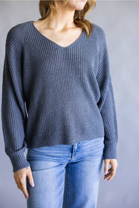 A woman wearing a slate blue ribbed knit sweater with a V-neck and long sleeves, paired with light blue jeans, shown from the neck down against a neutral gray background.