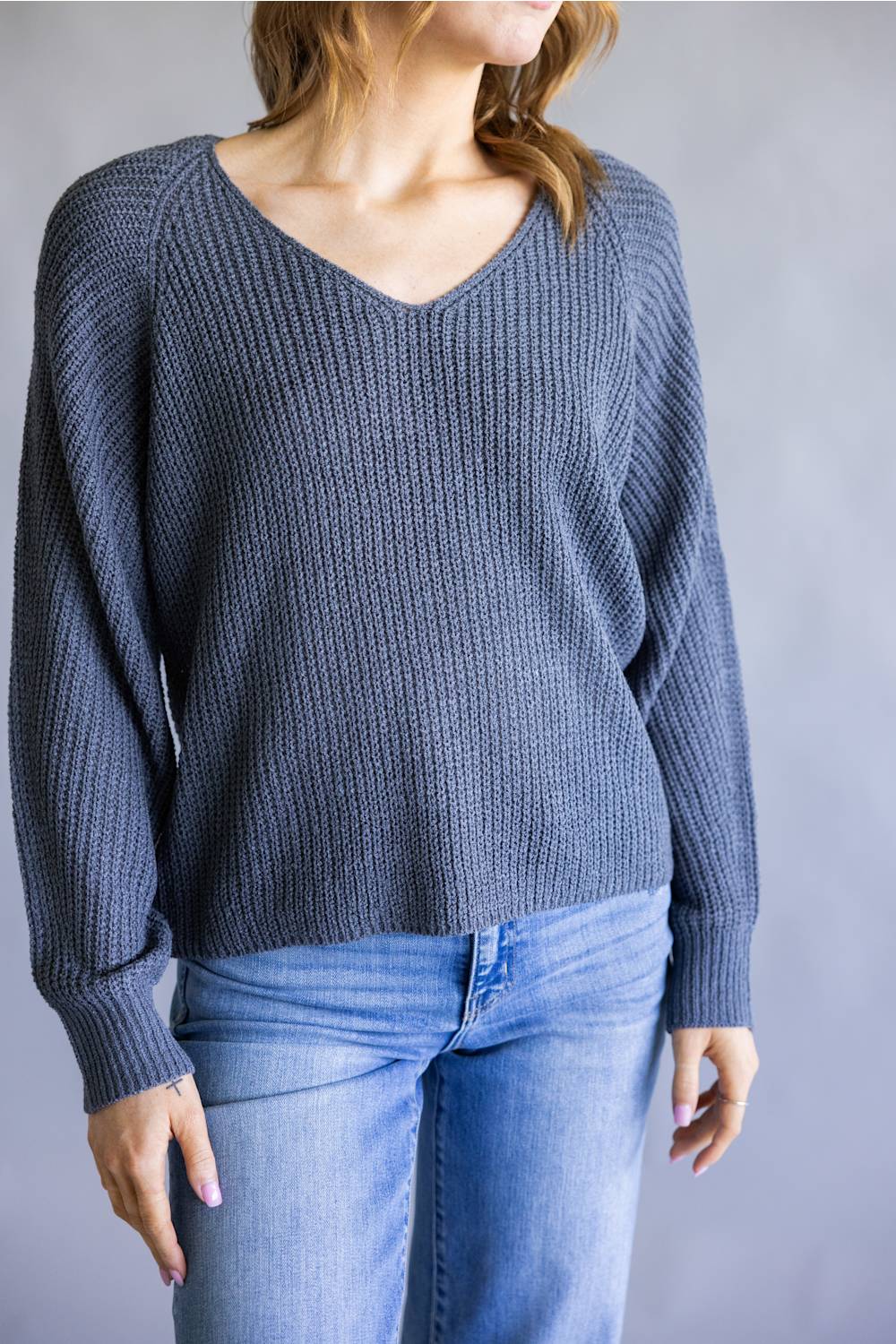 A woman wearing a slate blue ribbed knit sweater with a V-neck and long sleeves, paired with light blue jeans, shown from the neck down against a neutral gray background.
