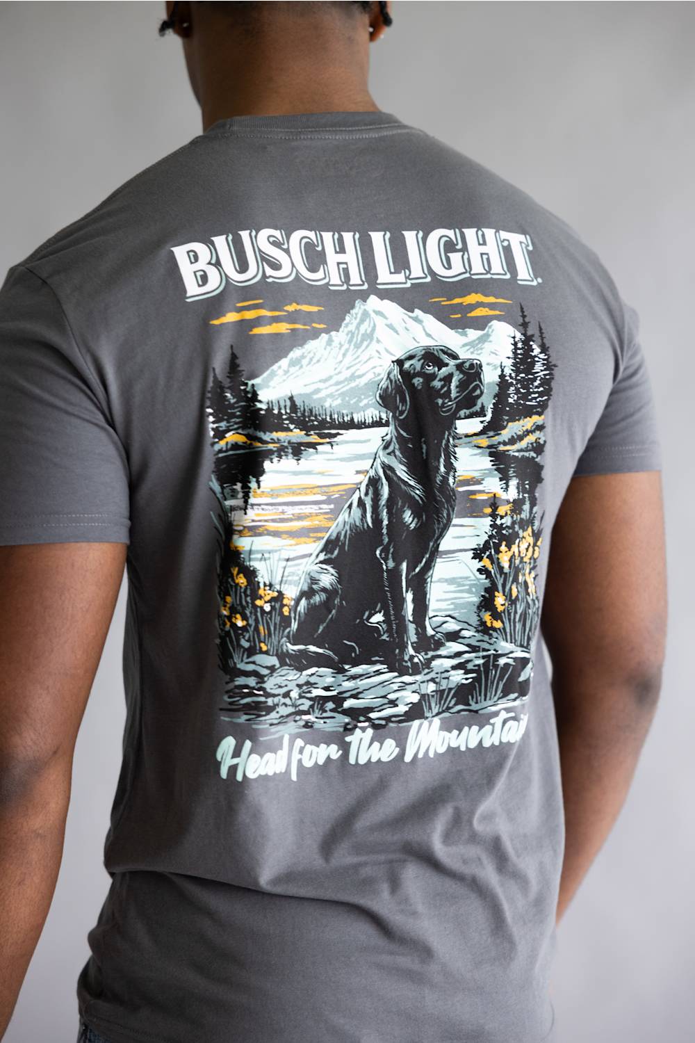 A man viewed from behind wearing a heather gray t-shirt with a large graphic print on the back. The graphic features the text 'BUSCH LIGHT' at the top and 'Head for the Mountains' at the bottom, surrounding an illustration of a black Labrador retriever sitting by a lake with a mountain in the background.