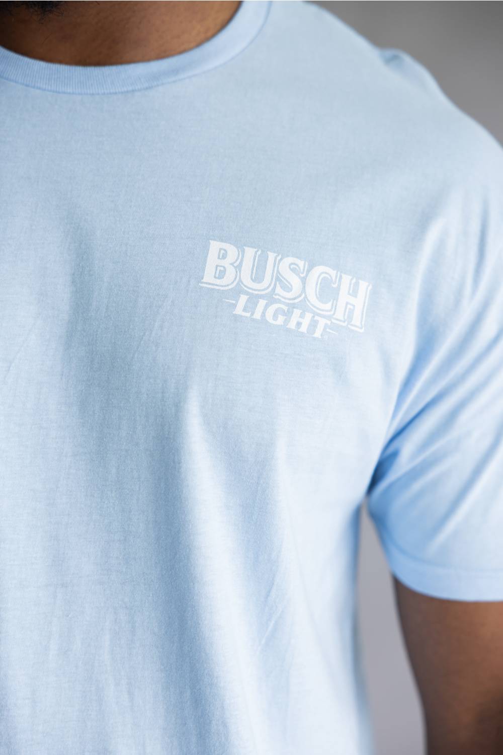 A close-up of a person wearing a light blue crewneck t-shirt with a white 'BUSCH LIGHT' logo printed on the chest.