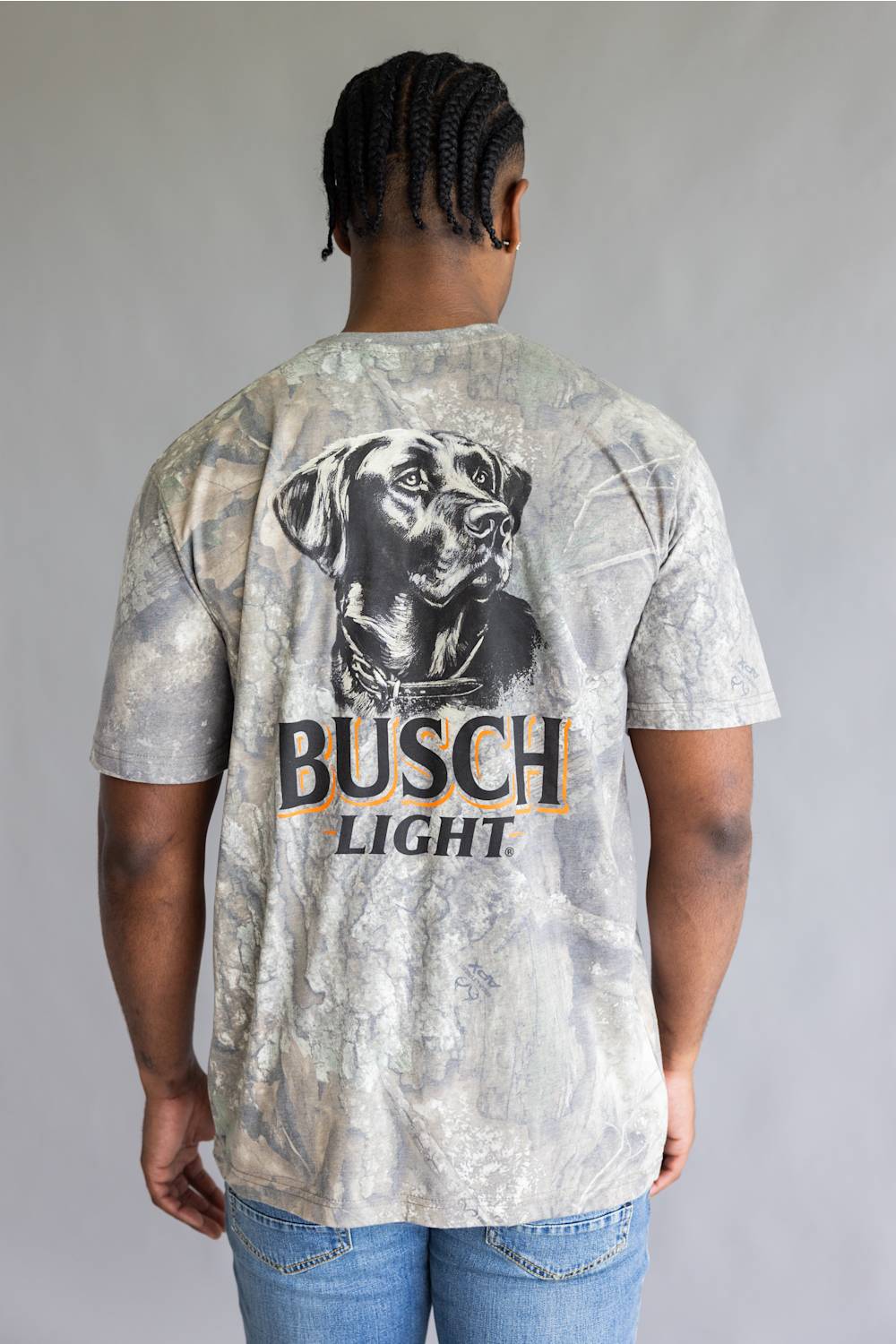 A man viewed from behind wearing a Busch Light t-shirt with a large black and white dog graphic and the brand name printed on the back, paired with blue jeans against a gray studio background.