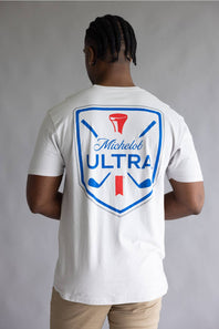 A man viewed from behind wearing a white Michelob ULTRA t-shirt with a large blue and red golf-themed graphic on the back, paired with khaki pants, against a neutral gray background.