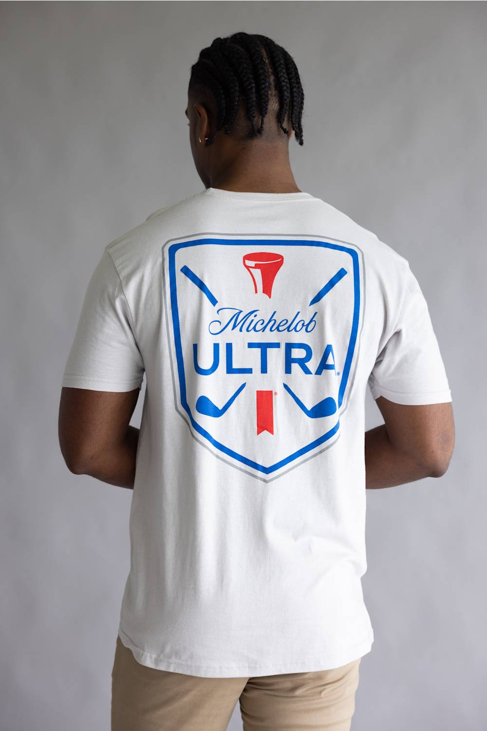 A man viewed from behind wearing a white Michelob ULTRA t-shirt with a large blue and red golf-themed graphic on the back, paired with khaki pants, against a neutral gray background.