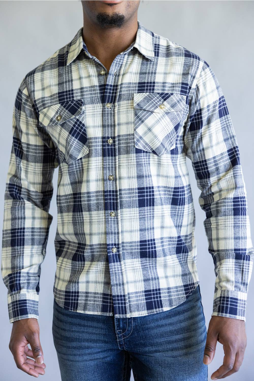 A man wearing a long-sleeve plaid button-up shirt in navy blue, cream, and white, paired with blue jeans, shown from the waist up against a neutral gray background.