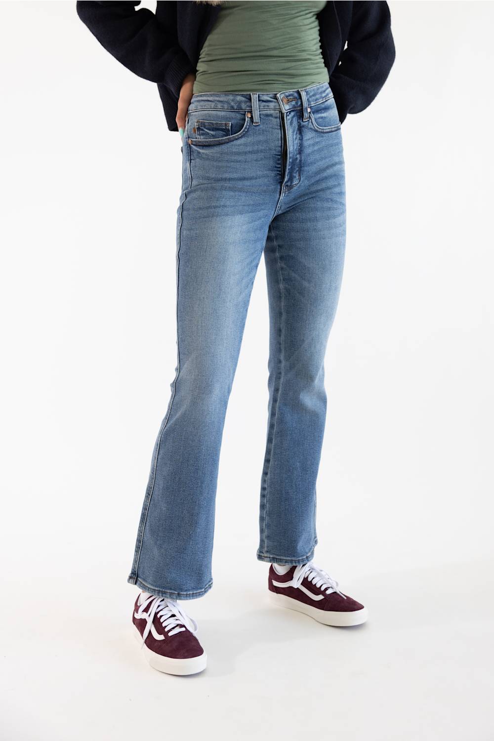 A person wearing light-wash blue flare jeans, a green top, a dark cardigan, and burgundy sneakers, standing against a plain white studio background.