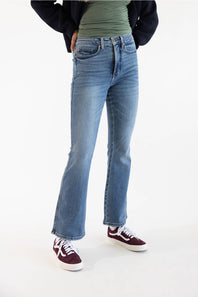A person wearing light-wash blue flare jeans, a green top, a dark cardigan, and burgundy sneakers, standing against a plain white studio background.