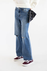 A person wearing medium-wash blue distressed flare jeans with ripped knees, styled with a white ribbed sweater, a navy patterned bandana, and burgundy sneakers against a plain white studio background.