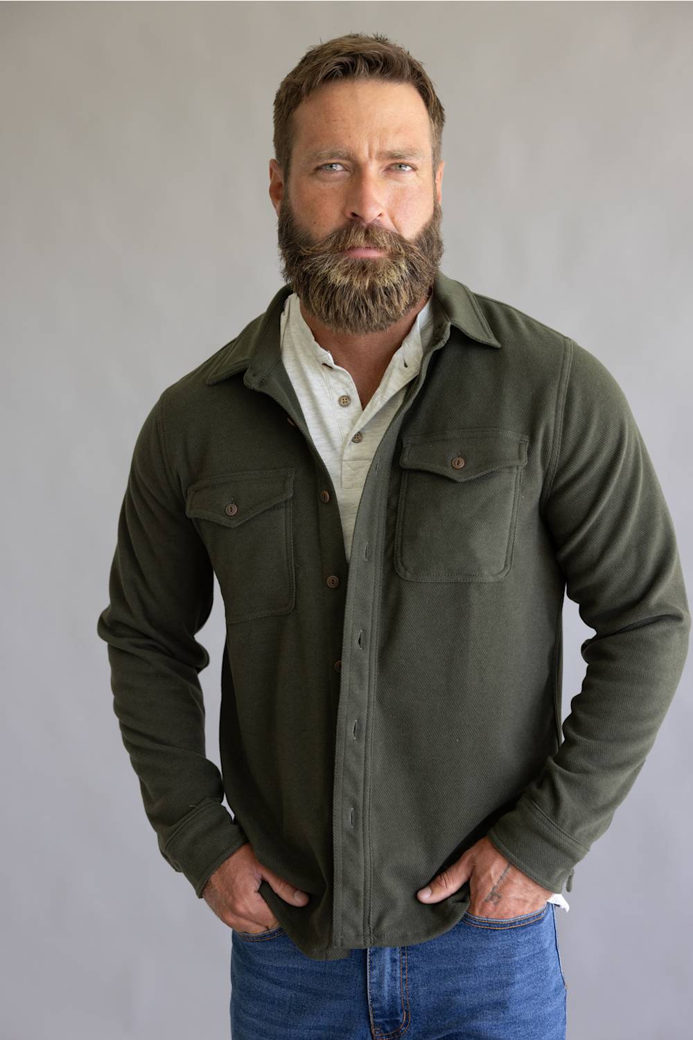 A man with a full beard wearing an olive green button-up shirt jacket over a white collared shirt and blue jeans, standing against a neutral gray studio background.