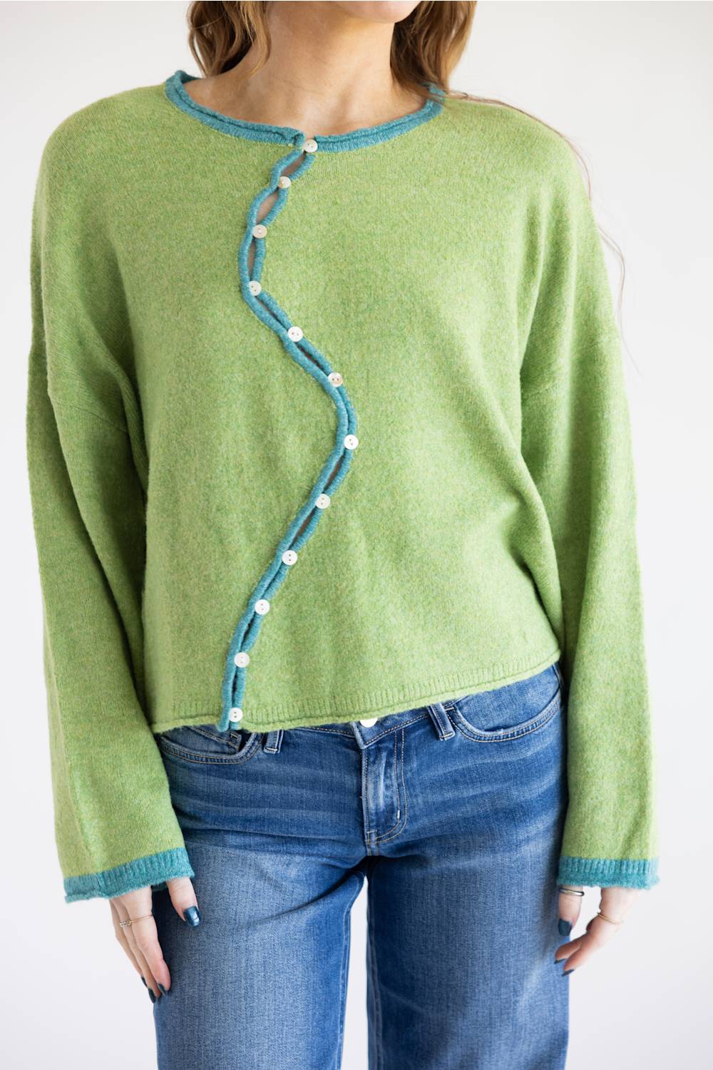 A woman wearing a light green knit sweater with a distinctive wavy front button closure and blue jeans, shown from the neck down against a plain white background.