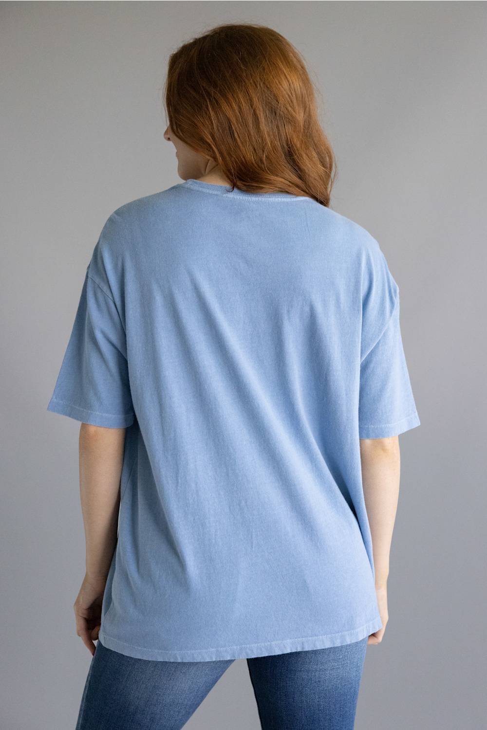 A woman with red hair is shown from the back wearing an oversized light blue t-shirt and blue jeans against a solid gray studio background.