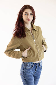 A woman with red hair wearing a cropped olive green utility jacket and blue jeans, posing against a plain white studio background.