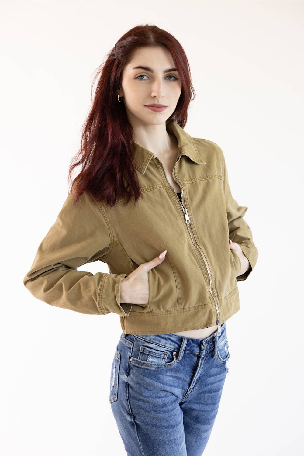 A woman with red hair wearing a cropped olive green utility jacket and blue jeans, posing against a plain white studio background.