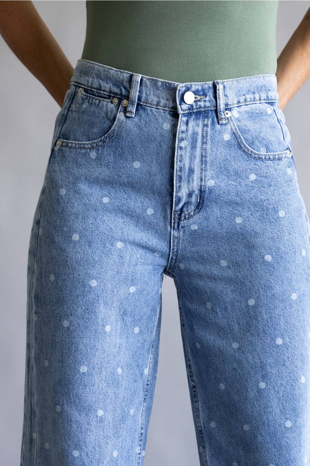 Close-up of a person wearing light blue denim jeans with a white polka dot pattern, paired with an olive green top against a neutral gray background.