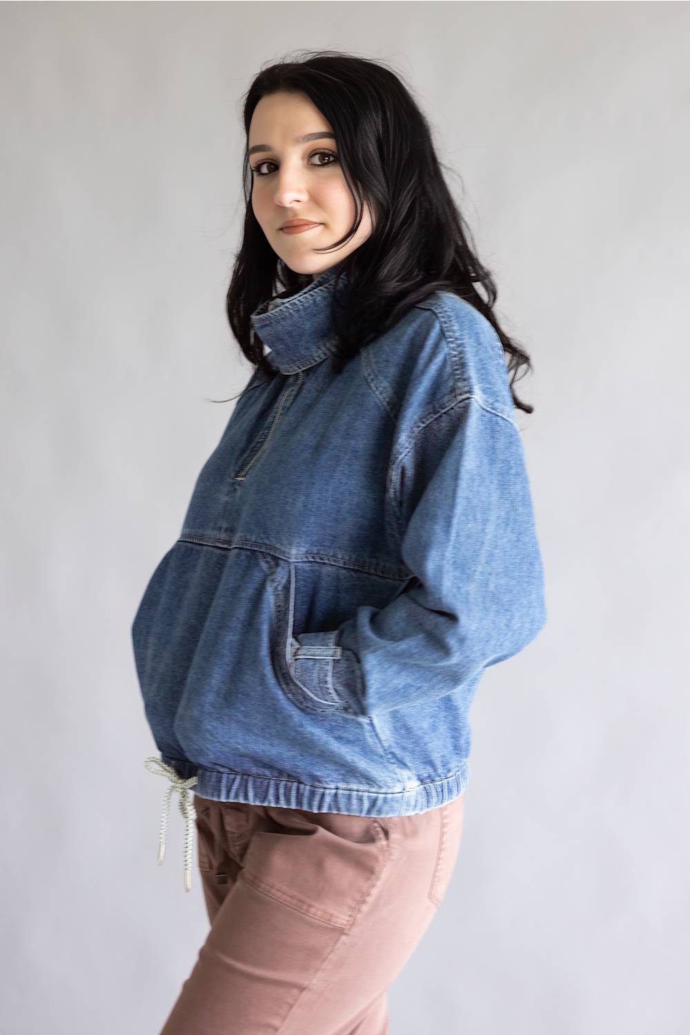 A woman wearing a blue denim half-zip jacket with a high collar and front pockets, paired with dusty rose pants, standing against a plain light gray background.