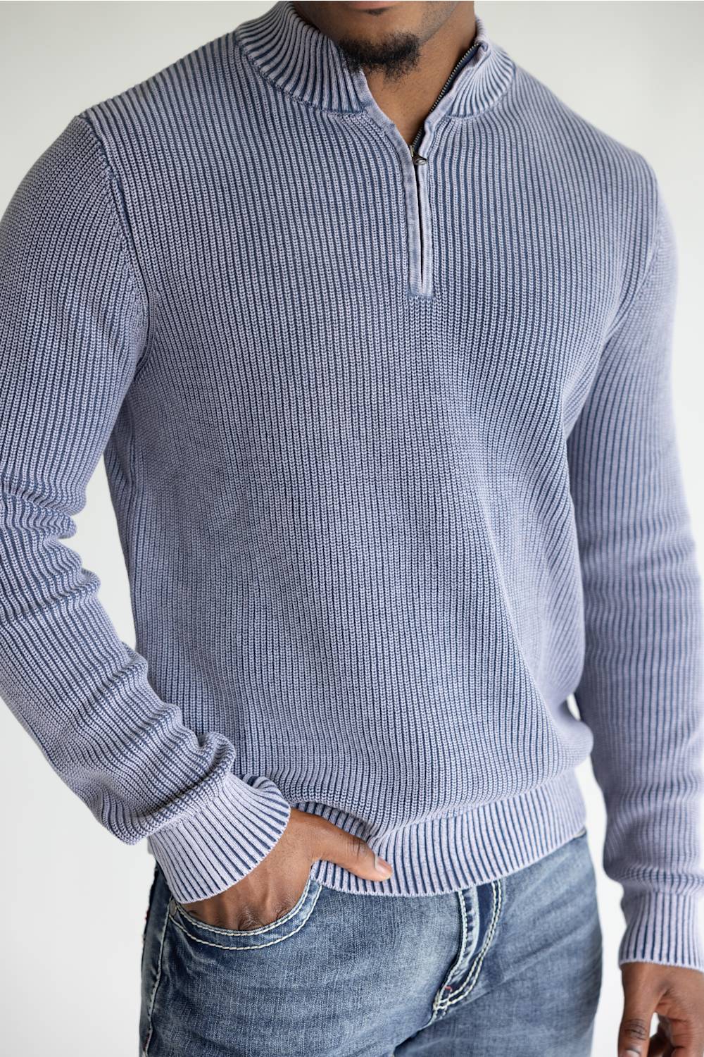 A man wearing a light blue and white vertically striped ribbed quarter-zip sweater with blue jeans, shown from the chest down against a plain white background.