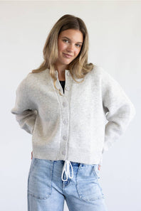 A woman modeling a light gray knit cardigan with a button-up front and drawstring detail, paired with light blue jeans against a plain white background.