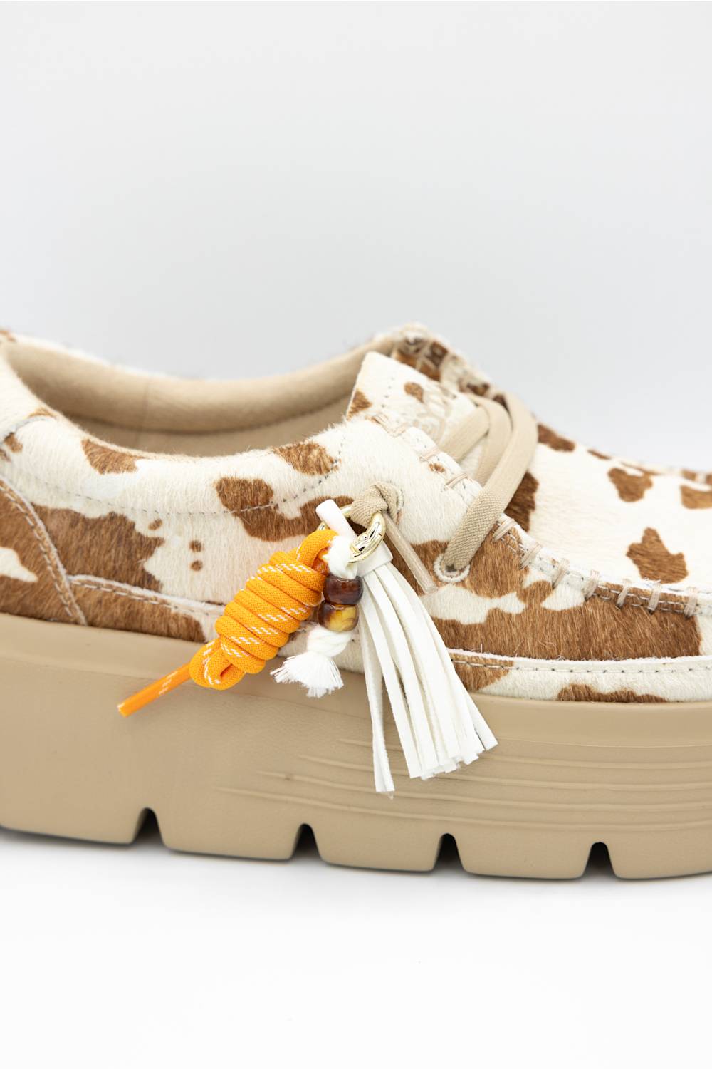 A close-up of a platform sneaker featuring a brown and cream cow-print pattern, beige sole, and decorative hanging tassels and an orange parrot charm.