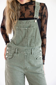 Close-up cropped view of Aaron & Amber Slouchy Denim Overalls for Women in Olive showing chest bib pocket details, silver adjustable strap hardware, three-button side closures, and front hand pockets paired over a black lace long-sleeve top.