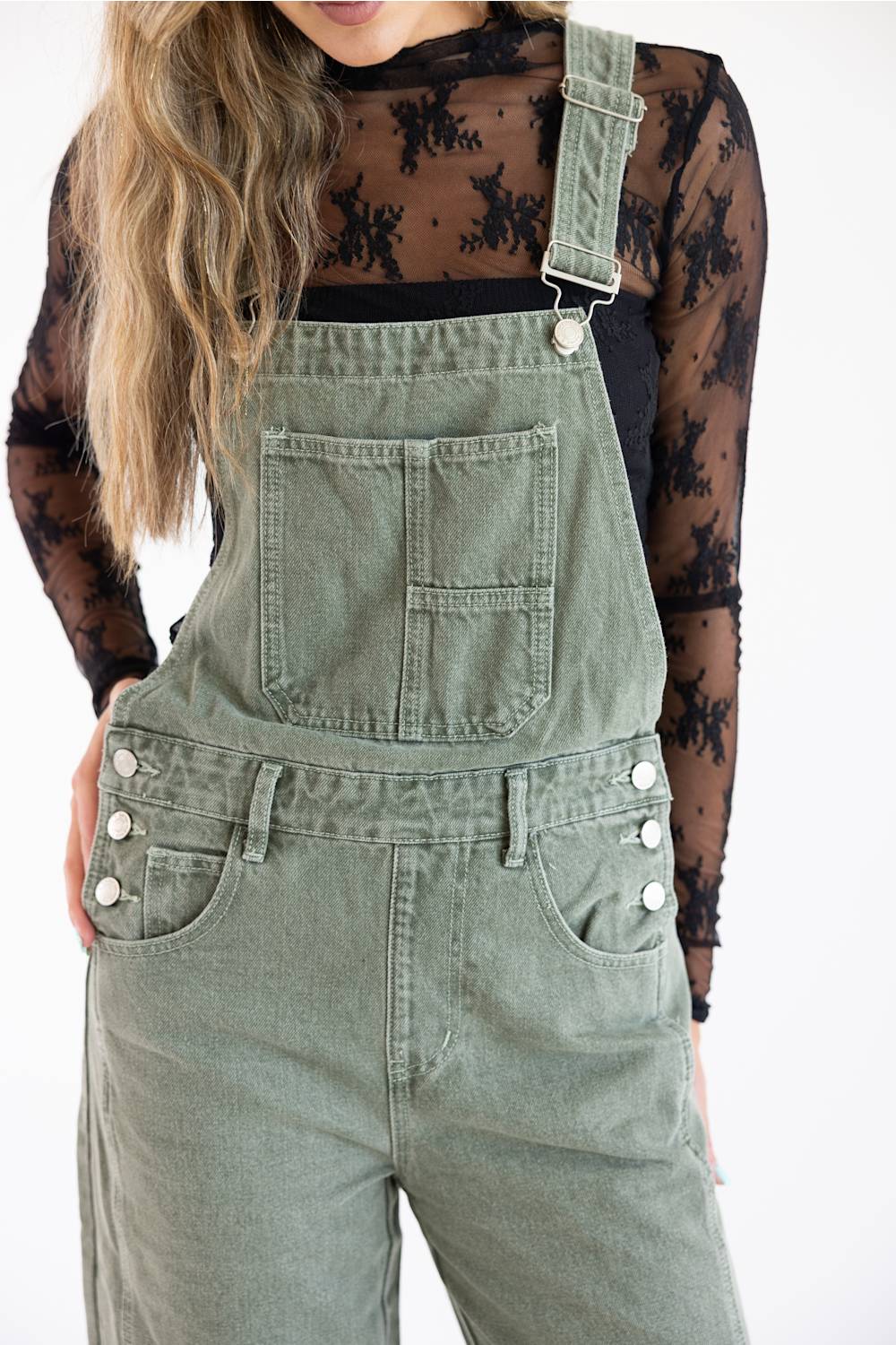 Close-up cropped view of Aaron & Amber Slouchy Denim Overalls for Women in Olive showing chest bib pocket details, silver adjustable strap hardware, three-button side closures, and front hand pockets paired over a black lace long-sleeve top.