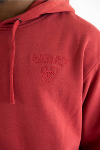 A close-up of a red hooded sweatshirt worn by a model, featuring an embossed Ariat logo on the chest.