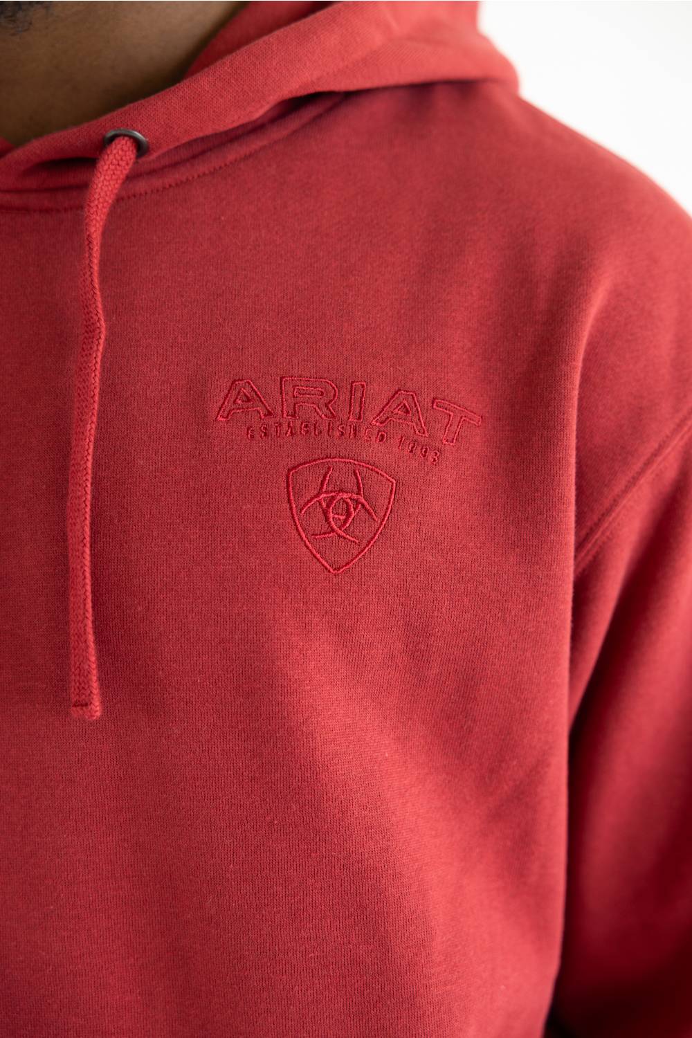 A close-up of a red hooded sweatshirt worn by a model, featuring an embossed Ariat logo on the chest.
