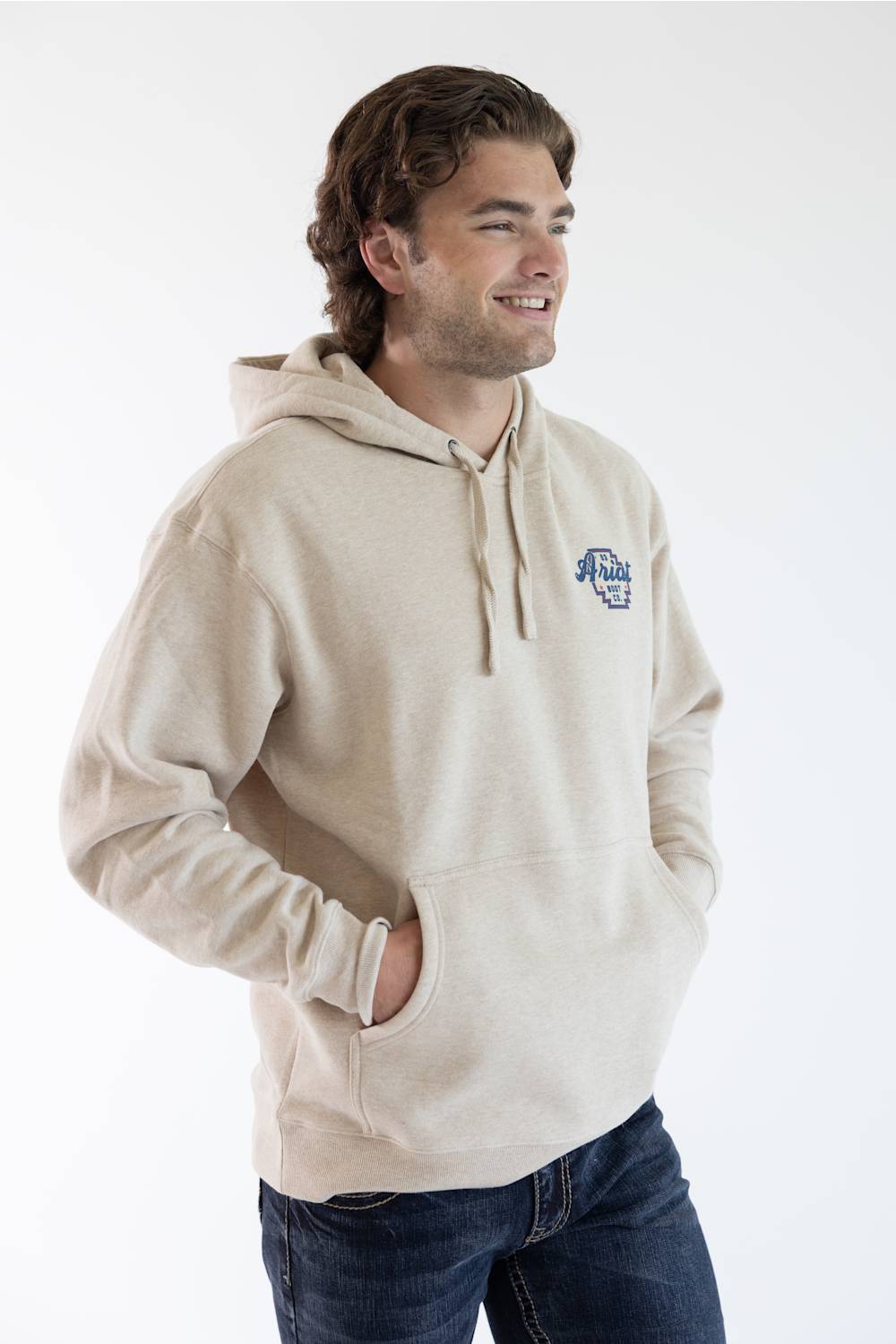 A man wearing a light beige pullover hoodie with a small blue and white graphic on the left chest, paired with dark blue jeans, photographed against a plain white studio background.