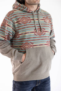 A man wearing a pullover hoodie with a colorful geometric pattern on the upper chest and hood, and a solid taupe-beige lower body and kangaroo pocket, paired with blue jeans against a white background.