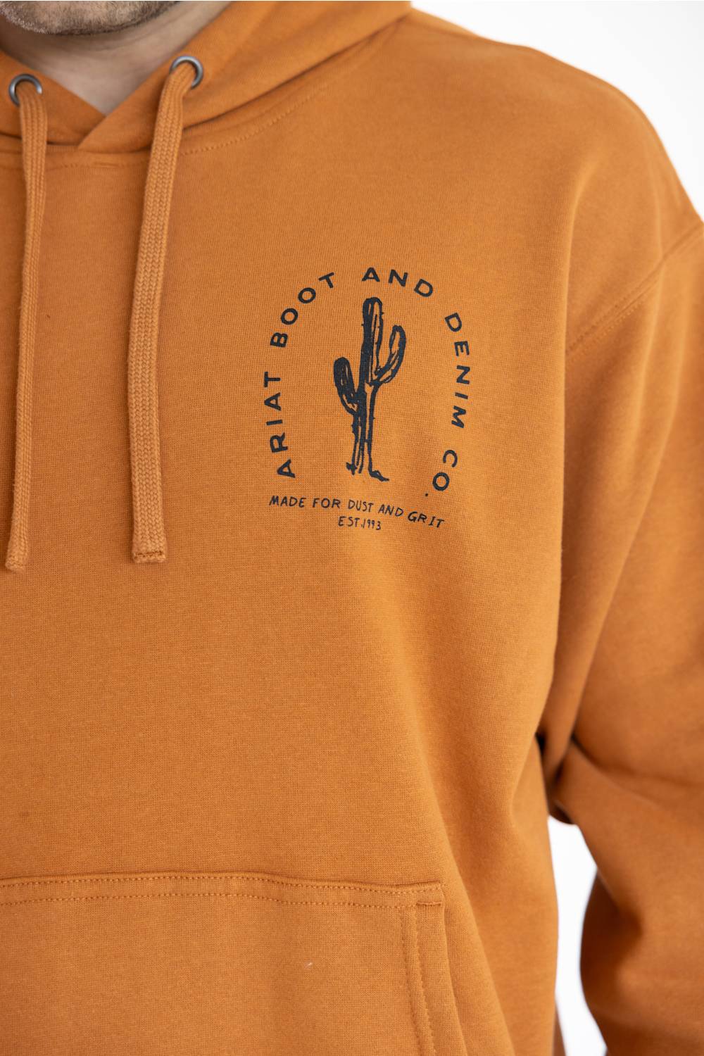 Close-up of a person wearing a mustard yellow hoodie with a black circular cactus graphic and the text 'ARIAT BOOT AND DENIM CO' printed on the chest.