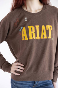 A woman with red hair wearing a brown crewneck sweatshirt with 'ARIAT' printed in yellow across the chest, paired with blue jeans, against a plain white background.