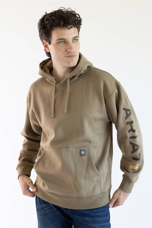 A man wearing a tan pullover hoodie with 'ARIAT' embroidered vertically in brown on the right sleeve and a small yellow logo patch near the cuff, paired with blue jeans against a plain white background.
