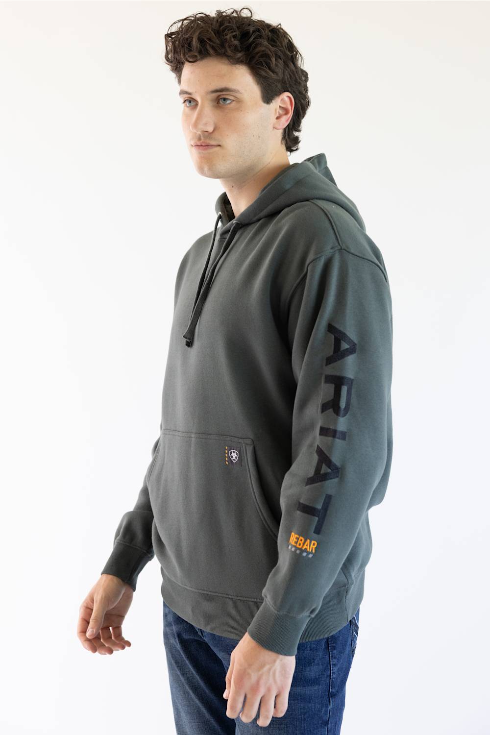 A man wearing a dark gray hoodie with 'ARIAT' printed vertically in black on the right sleeve and a small orange logo near the pocket, paired with blue jeans against a plain white background.