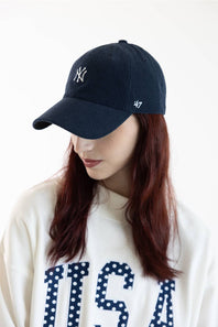A woman wearing a dark navy blue corduroy baseball cap with a white embroidered New York Yankees logo on the front and a small '47' brand logo on the side, paired with a cream sweatshirt.