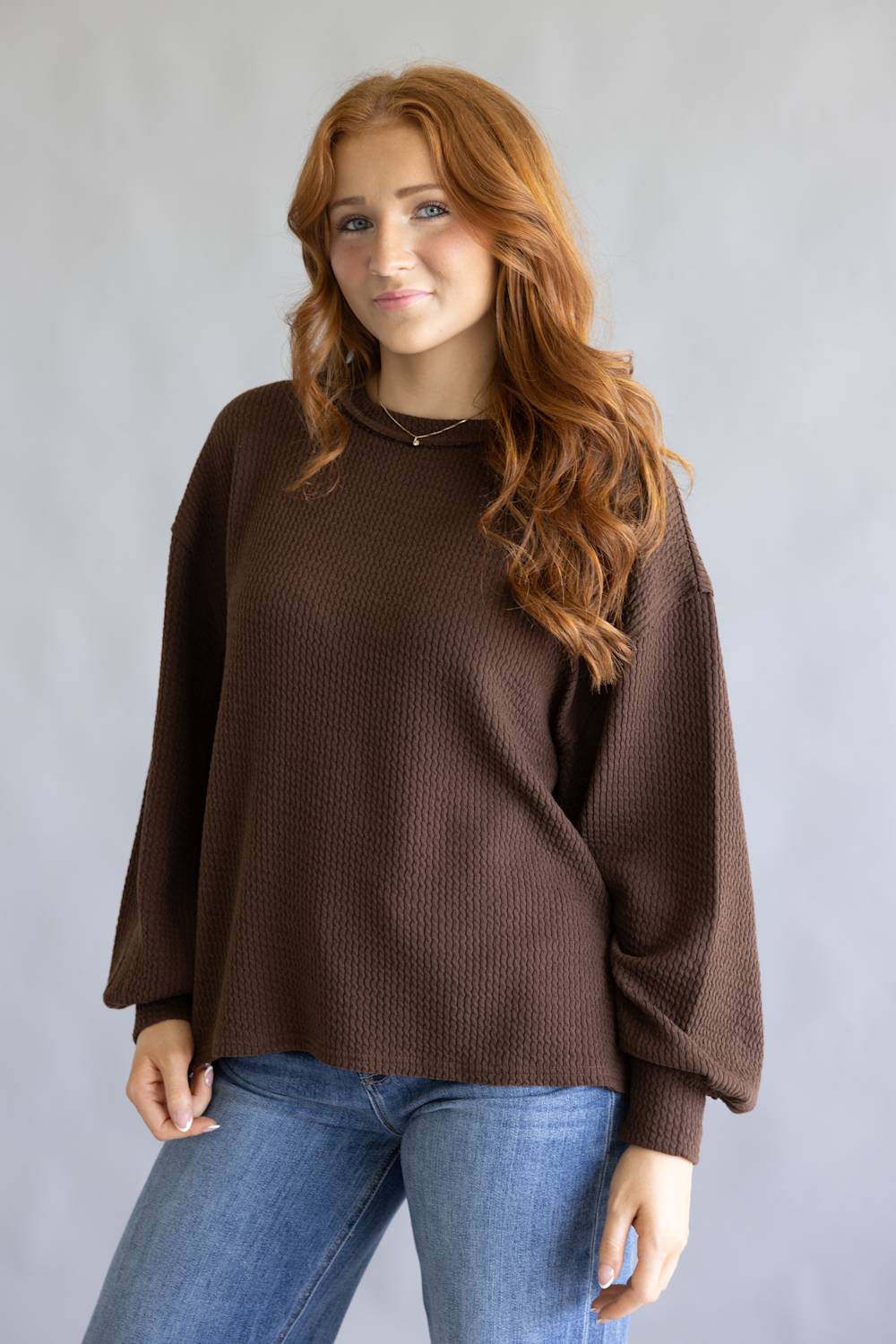 A woman with long red hair wearing a brown waffle-knit long-sleeve sweater and blue jeans, standing against a light gray studio background.
