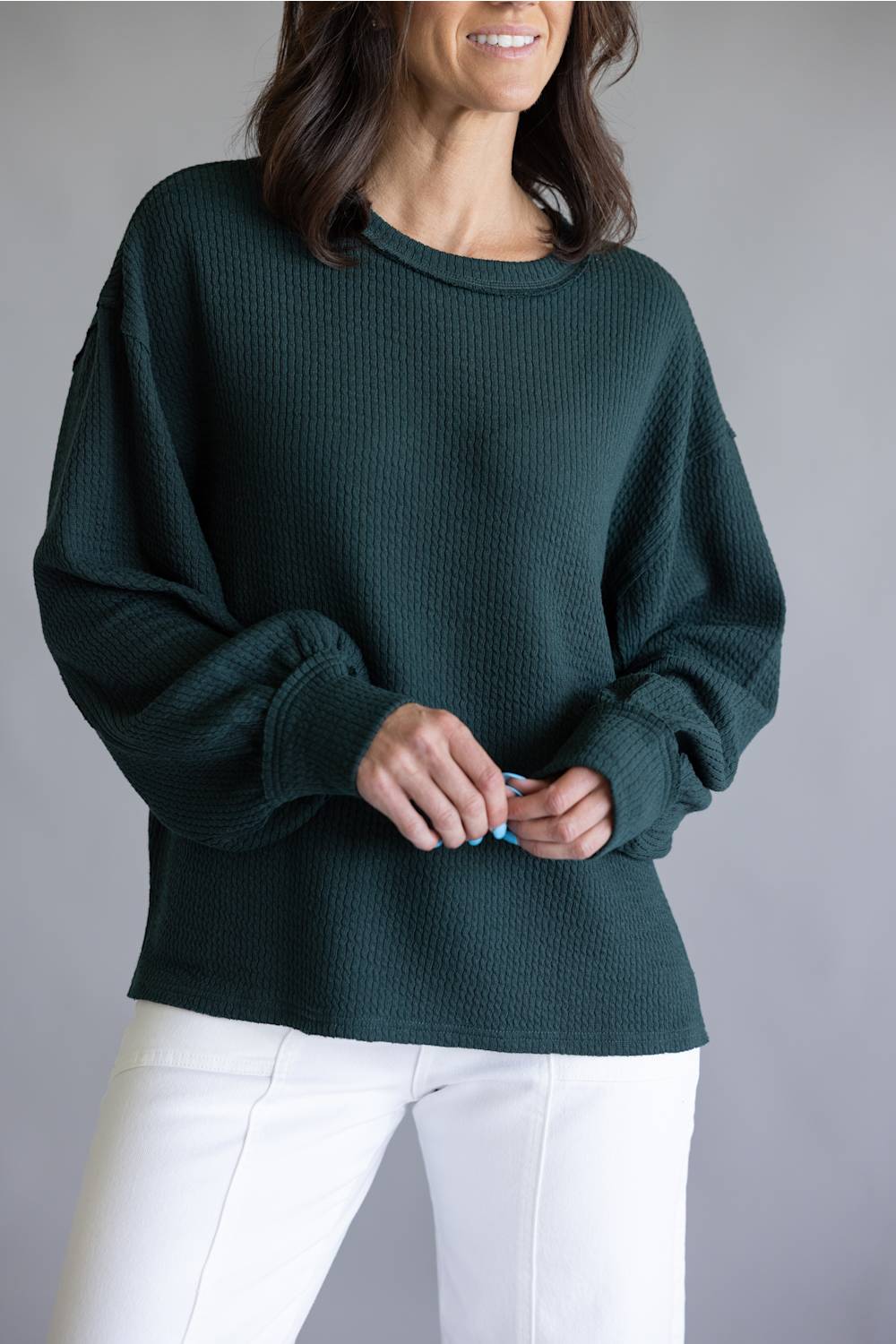 A woman wearing a dark forest green waffle-knit sweater with balloon sleeves and white pants, standing against a neutral gray studio background.