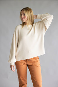 A woman modeling a cream-colored, textured knit sweater with wide three-quarter sleeves, paired with rust-orange pants, against a solid gray studio background.