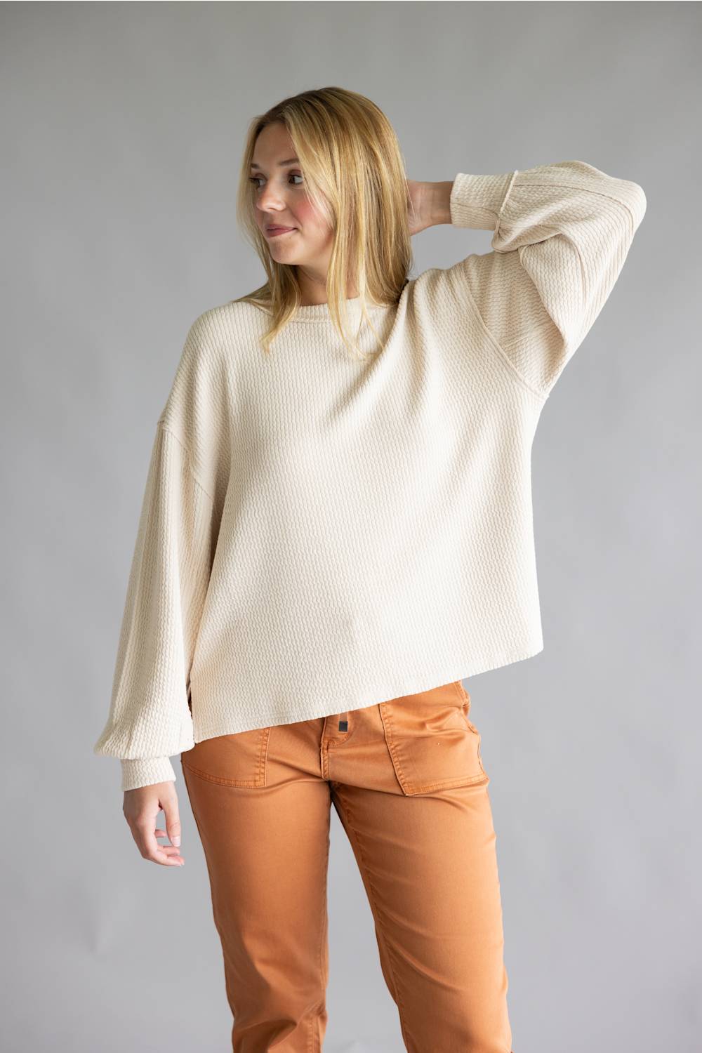 A woman modeling a cream-colored, textured knit sweater with wide three-quarter sleeves, paired with rust-orange pants, against a solid gray studio background.