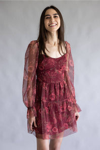 A woman wearing a tiered burgundy floral dress with sheer long sleeves, standing against a neutral gray studio background.