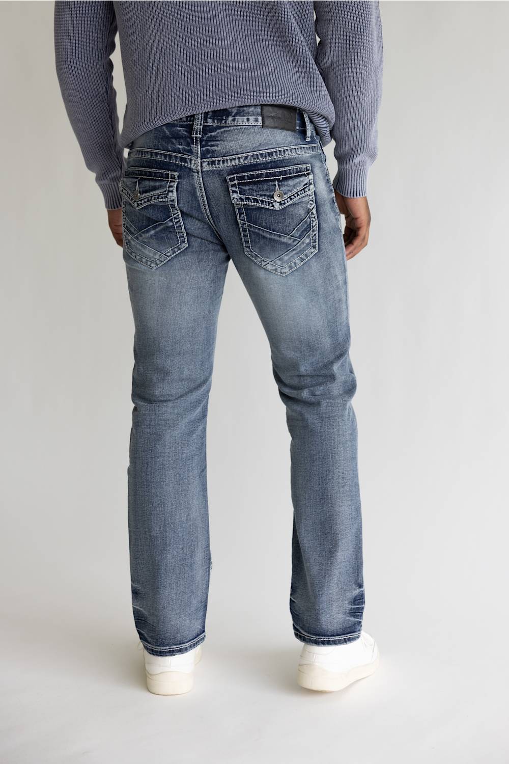 Rear view of a person wearing light blue denim jeans with a ribbed gray sweater and white sneakers, standing against a plain light gray studio background.