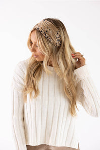 A woman with long blonde hair wearing a beige patterned headband and a cream ribbed knit sweater, posing against a solid white background.