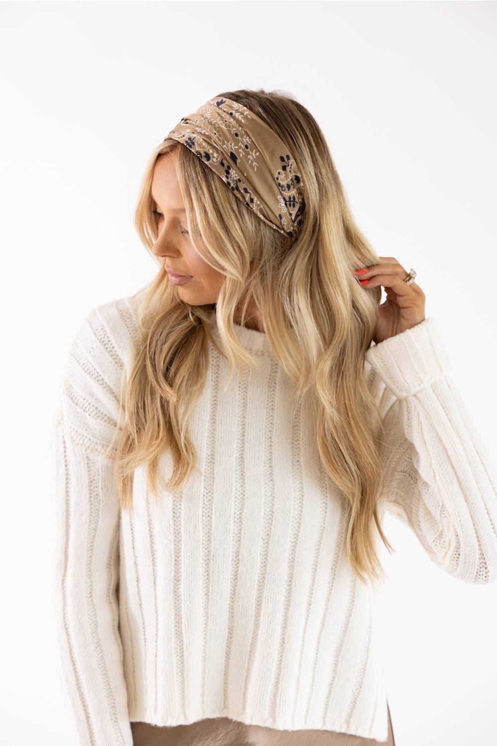 A woman with long blonde hair wearing a beige patterned headband and a cream ribbed knit sweater, posing against a solid white background.