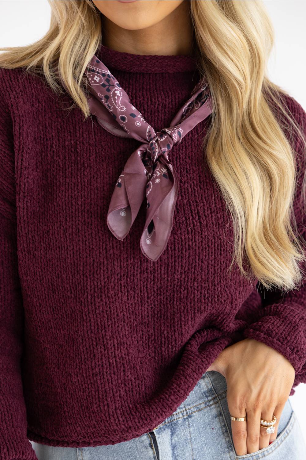 A close-up of a woman wearing a chunky knit burgundy sweater and a mauve paisley neck scarf tied in a knot at the collar.